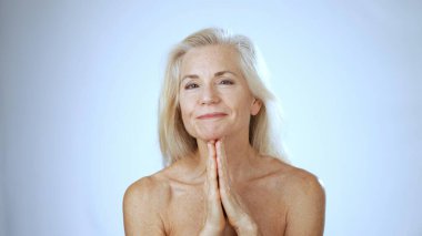 Olgun bir kadın, basit bir beyaz arka plana karşı barışçıl bir jest yaparak ellerini birleştirip mutlu bir şekilde bir kaplıcada dinlenir..