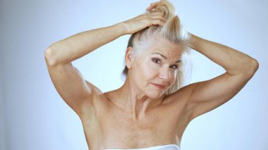 Kaplıcadaki olgun bir kadın neşeli bir ifade sergilerken saçlarını mutlu bir şekilde tarar. Arka plan parlak ve beyaz, canlandırıcı atmosfere katkı sağlıyor..