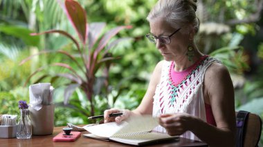 Orta yaşlı bir kadın enerjik bir kafede oturur ve defterine odaklanır. Bereketli yeşilliklerle çevrili, seyahatleri sırasında işi ve rahatlığı dengeliyor..