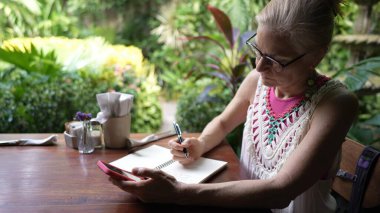 Orta yaşlarındaki bir kadın kafeterya masasında oturmuş, telefonuna odaklanmış, etrafı canlı bitkiler ve huzurlu bir atmosferle çevrili bir defterden notlar alıyor..