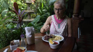 Olgun bir kadın, yemyeşil yemyeşil bir kafede besleyici bir yemeğin tadını çıkarıyor. Tatili boyunca işi ve rahatlığı dengeliyor..