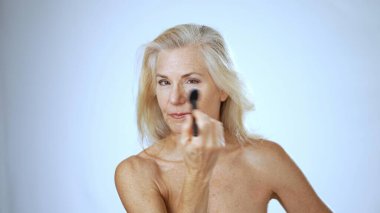 Olgun bir kadın, sakin bir spa ortamında makyaj yaparken gülümser. Parlak arka plan onun mutluluğunu arttırıyor, neşeli bir atmosfer yaratıyor..