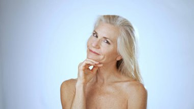 Uzun, dalgalı saçları olan olgun bir kadın neşeli bir şekilde gülümserken berrak, sakin bir spa ortamında rahatlıyor. İfadesi mutluluk ve huzur veriyor..