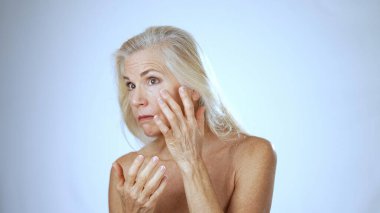 Olgun bir kadın spa tedavisi sırasında yüzüne krem sürerken mutlu bir şekilde gülümser. Arka plan sakin bir beyaz, neşe dolu deneyimini arttırıyor..