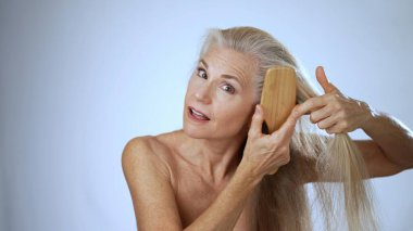 Olgun bir kadın, uzun ve güzel saçlarını tararken huzurlu bir spa ortamında mutlu bir şekilde gülümser. Yumuşak beyaz bir arka plan, neşe ve rahatlama saçar..
