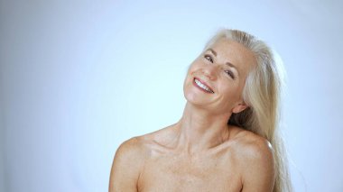 Olgun bir kadın huzurlu bir spa ortamında mutlu bir şekilde oturur, yüzünde sıcak bir gülümsemeyle mutluluk saçar. Onun uzun, dalgalı saçları onun rahat görünümüne ekleniyor..