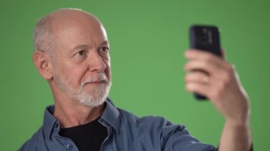 Olgun bir adam gülümser ve canlı yeşil bir arkaplan karşısında selfie çekerken başparmağını kaldırır. Neşeli tavrını ve gevşemiş havasını gösterir..