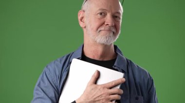 Olgun, orta yaşlı bir adam gözlükleriyle ayakta duruyor, göğsüne yakın bir tablet tutuyor parlak yeşil bir ekrana karşı. Rahat ve cana yakın görünüyor..