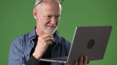 Olgun bir adam dizüstü bilgisayarına odaklanır, yeşil bir ekranın önünde oturur. Konsantrasyonunu göstermek için önemli bir işle meşgul gibi görünüyor..