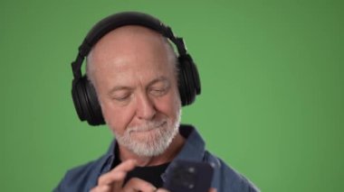 Neşeli bir ifadesi olan orta yaşlı bir adam yeşil arka planda akıllı telefonuyla iletişim kurarken kulaklıkla müzik dinler..