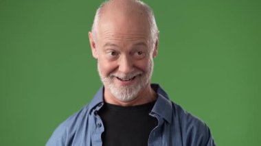 Sakallı orta yaşlı bir adam neşeli bir yüz ifadesi sergilerken, parlak yeşil bir perde arkasında neşeli bir jest yaparak, davetkar bir atmosfer yaratıyor..