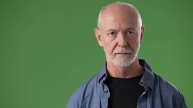 Orta yaşlı bir adam elini kaldırarak seyirciyi selamlıyor. Kendine güveni tam ve yeşil ekran arka planına karşı..