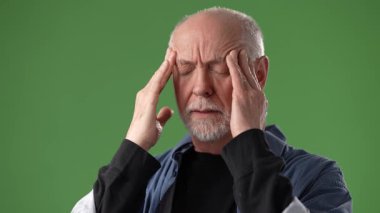 Gri saçlı orta yaşlı bir adam stres yaşıyor gibi görünüyor. Şakaklarını yeşil ekran arka planı olan bir stüdyoda tutuyor..