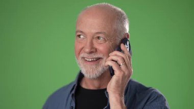 Olgun bir adam telefonuyla konuşurken gülümsüyor, dostça bir tavır sergiliyor. Gri bir sakalı var ve kot gömlek giyiyor. Arkaplan canlı yeşil.