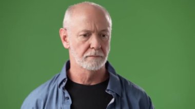 Ciddi bir ifadeye sahip orta yaşlı bir adam, parlak yeşil bir ekrana karşı duruyor ve görünüşünü ve kıyafetlerini vurguluyor. Gümüş rengi saçları ve sakalı var..