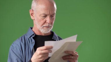 Gri sakallı olgun bir adam, açık yeşil bir ekranın önünde dururken odaklanmış bir ifadeyle belgeler okur..