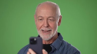 Olgun bir adam, parlak yeşil bir ekrana karşı akıllı telefon kullanarak gülümser ve el sallar. Neşeli ve bağlı görünüyor, sanal bir sohbetten hoşlanıyor..