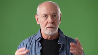 Gri saçlı ve sakallı olgun bir adam sürpriz bir şekilde, sıradan bir kıyafet giyiyor. Canlı yeşil bir arkaplana karşı duruyor ve ifadesine net bir şekilde odaklanıyor..