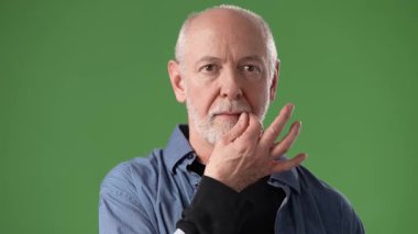 Orta yaşlı, sakallı bir adam elini yeşil ekran arka planı olan bir stüdyoda nazikçe dinlenen izleyiciye bakar..