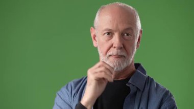 Olgun, orta yaşlı bir adam, hafif bir gülümseme ile kendine güvenerek duruyor, elini parlak yeşil bir perde arkasında işaret ediyor..