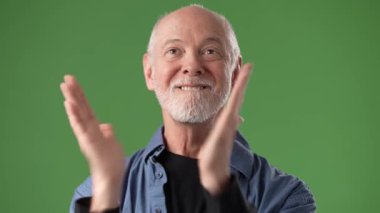 Gülümseyen orta yaşlı bir adam canlı yeşil bir ekranın önünde neşeli tavırlarını sergiliyor. Koyu renk bir bluzun üzerine sıradan bir gömlek giyer..
