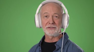 Olgun bir adam, canlı yeşil bir ekrana karşı poz verirken kulaklık takar ve gülümser. Stüdyo ortamında sanal etkileşime ya da kayıtlara hazırdır..