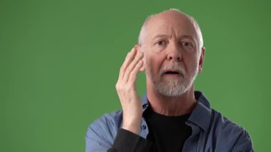 Sakallı olgun bir adam, canlı yeşil bir zemine karşı elini kaldırırken geniş bir gülümseme ile gülümseyerek dostça bir ifade sergiliyor..