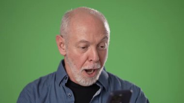 Sakallı olgun bir adam canlı yeşil bir ekrana karşı akıllı bir telefon tutarken heyecan gösterir. Onun ifadesi şaşkınlık ve mutluluk taşıyor..
