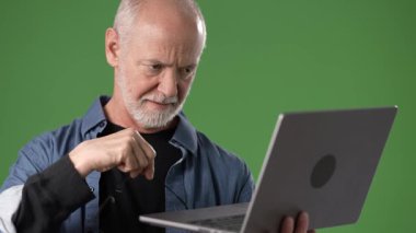 Gri saçlı ve gözlüklü orta yaşlı bir adam dikkatle dizüstü bilgisayar kullanıyor. Ekrana yoğunlaşıyor gibi görünüyor, yeşil ekran arka planına göre ayarlı..