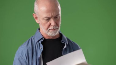 Olgun bir adam, bir mektup okurken şaşırmış bir şekilde yeşil bir ekrana karşı duruyor. Sıradan giyinmiş ve ellerindeki içeriğe odaklanmış görünüyor..