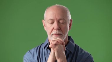 Sakallı, olgun, orta yaşlı bir adam stüdyo ortamında parlak yeşil bir perdeye dayanan düşünce ya da dua jesti ile ellerini kaldırır..