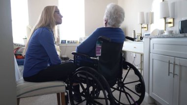 Bir kadın sandalyeye oturur ve yaşlı annesiyle evde tekerlekli sandalyede samimi bir konuşma yapar. Onların bağları günlük hayatta sevgi ve ilgiyi yansıtır..