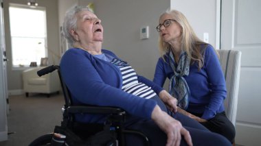 Kızı evde duygusal destek sağlarken 90 'larında yaşlı bir kadın tekerlekli sandalyede oturuyor. Sıcak bir ortamda bir anlık birlikteliği paylaşıyorlar..