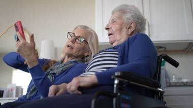 Kapalı bir ortamda, tekerlekli sandalyedeki yaşlı bir kadın akıllı telefon aracılığıyla kızıyla iletişim kuruyor, gülümsemelerini ve güzel anılarını paylaşıyor..