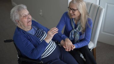 90 'lı yaşlarda bir kadın tekerlekli sandalyede oturuyor, kızıyla canlı bir şekilde konuşuyor, evini ziyaret ederken sıcak ve şefkatli bir ortam yaratıyor..