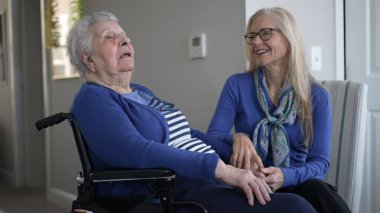 Ev ortamında, bir kız, yaşlı annesinin elini sevgiyle tutar. Hem gülümser, hem de evde eğlenir..