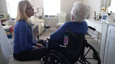 Sıcak, kapalı bir mekanda, bir kadın yaşlı annesine yakın bir tekerlekli sandalyede oturuyor, aile ziyareti sırasında bir anlık ilgi ve bağlantı paylaşıyor..