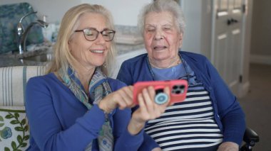 Tekerlekli sandalyedeki yaşlı bir kadın, kızıyla akıllı telefon kullanırken gülümsüyor..