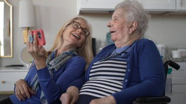 Tekerlekli sandalyedeki yaşlı bir kadın, kızı ona akıllı bir telefon gösterirken gülümsüyor. Sıcak bir evde sevgi ve beraberlik anları yakalıyor..