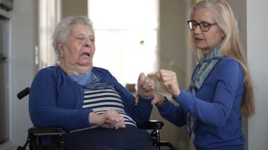 Bir kız, evde yaşlı annesine tekerlekli sandalyede yardım ediyor. Ev ortamındaki günlük aktivitelerinde sevgi ve ilgi gösteriyor..