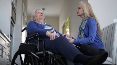 Tekerlekli sandalyedeki yaşlı bir kadın sıcak bir ev ziyareti sırasında kızından sevgi dolu destek alıyor. Birlikte sıcak bir anı paylaşıyorlar. Parlak, davetkar bir yerde..