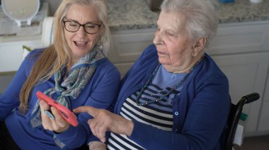 Tekerlekli sandalyedeki yaşlı bir kadın kızıyla görüntülü sohbet etmekten zevk alıyor. Sıcak mutfaklarında gülümseme ve anılarını paylaşıyor. Sevgi ve desteği vurguluyor..