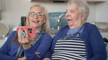 Tekerlekli sandalyedeki yaşlı bir kadın kızıyla neşe içinde iletişim kuruyor, akıllı bir telefondan hatıralarını paylaşıyor samimi bir ev ziyareti sırasında..