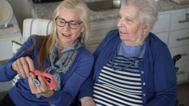 Kapalı bir ortamda, 60 'lı yaşlardaki bir kadın yaşlı annesinin fotoğraflarını akıllı bir telefondan gösteriyor, ziyaretleri sırasında sevgi ve bağlantı anlarını yakalıyor..
