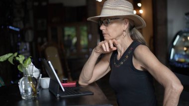 Olgun bir kadın bir kafede oturur, çenesini elinde dinlendirirken tabletine odaklanır. Akıllı telefon ve kahve fincanı yakınlarda dijital göçebe yaşam tarzını sergiliyor..