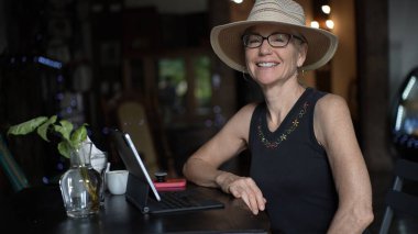 Olgun bir kadın, tableti ve bilgisayarıyla uğraşırken kafeterya masasında gülümsüyor. Taze yeşillik ve sıcak atmosfere bir kahve fincanı ilave edilir..