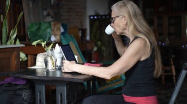 Olgun bir kadın kahve masasında oturur, kahvesini yudumlar ve telefonuyla birden fazla iş yaparken tablet kullanır. Rahat atmosfer onun dijital göçebe yaşam tarzını tamamlıyor..