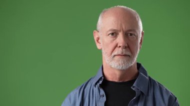 Sakallı, olgun bir adam stüdyoda sakin bir ifade sergiliyor. Siyah bir tişörtün üzerine günlük mavi bir gömlek giyer. Çekime hazırdır..