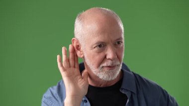 Orta yaşlı, sakallı ve kısa saçlı bir adam eli kulağında, canlı yeşil bir arka plana karşı özenli bir ifade sergiliyor..