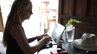 Olgun bir kadın kafeterya masasında oturup bir fincan kahvenin tadını çıkarırken tabletine odaklanır. Dijital göçebe yaşam tarzı çalışma alanından belli oluyor..